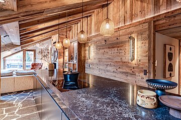 Rustic modern chalet interior featuring a dark marble kitchen island, wood-paneled walls, and warm woven pendant lights.