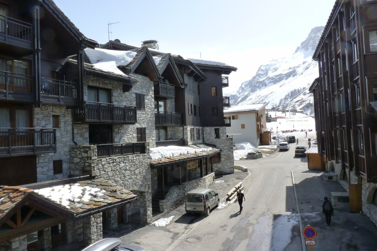Alpine village street scene with snow-dusted stone chalets, cars, people, and snowy mountains in the background.