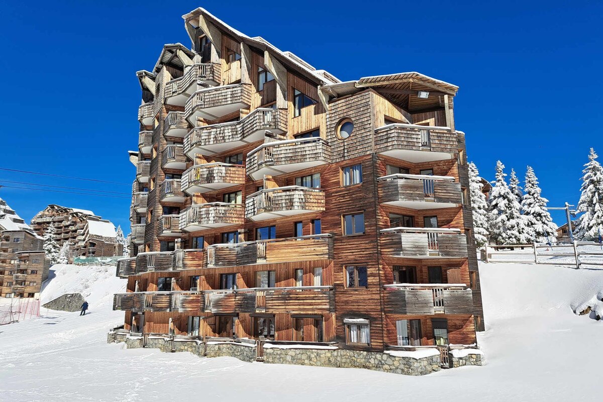 A large wooden ski resort building with many balconies, covered in snow under a bright blue sky. Snowy trees and a skier are visible.