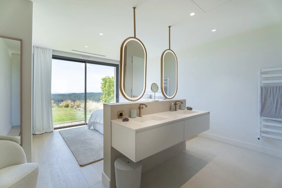 Modern open-concept bedroom and bathroom with dual sinks, illuminated mirrors, and a large window overlooking a scenic landscape.