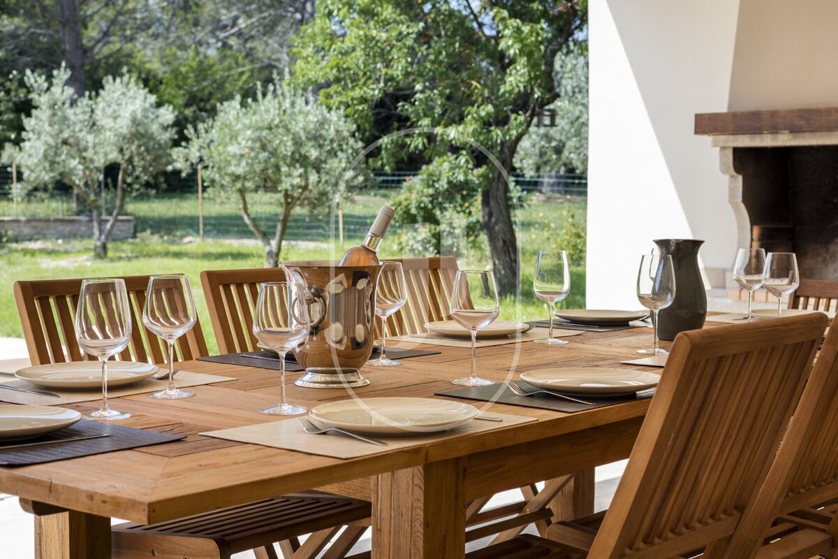 An elegant outdoor dining setup on a wooden table, featuring wine, glasses, and plates, against a scenic backdrop of trees and a rustic fireplace.