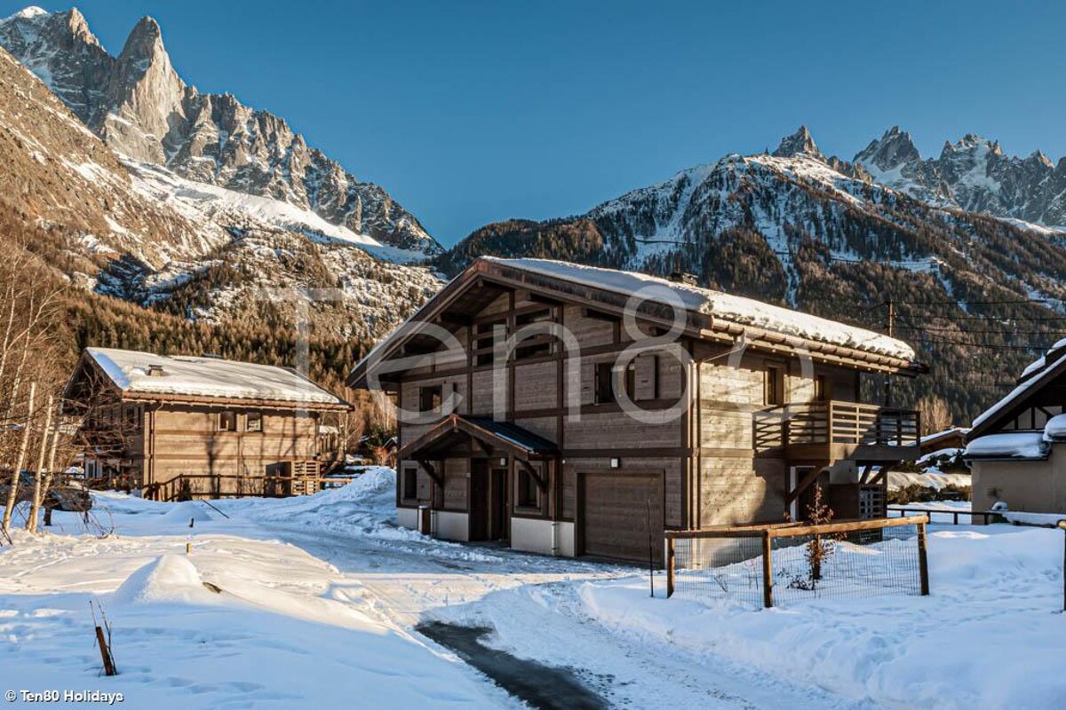 Snow-covered wooden chalets sit amidst a pristine winter landscape, flanked by towering, snow-capped mountains under a clear blue sky.