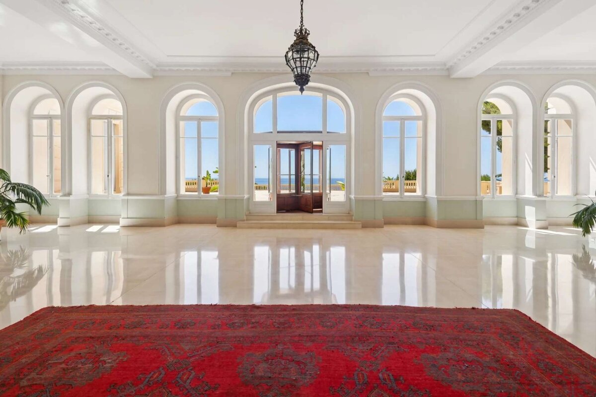 An opulent hall with arched windows, a red rug, and reflective marble floors. A central doorway reveals a stunning ocean view, illuminated by a hanging lantern.