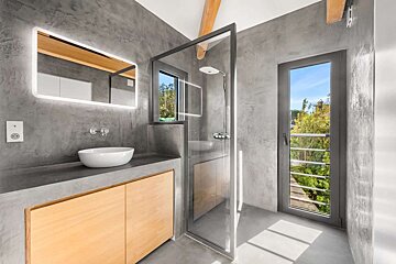 Modern, minimalist bathroom with gray concrete walls, wooden vanity, white vessel sink, backlit mirror, glass shower, and a large window overlooking greenery.