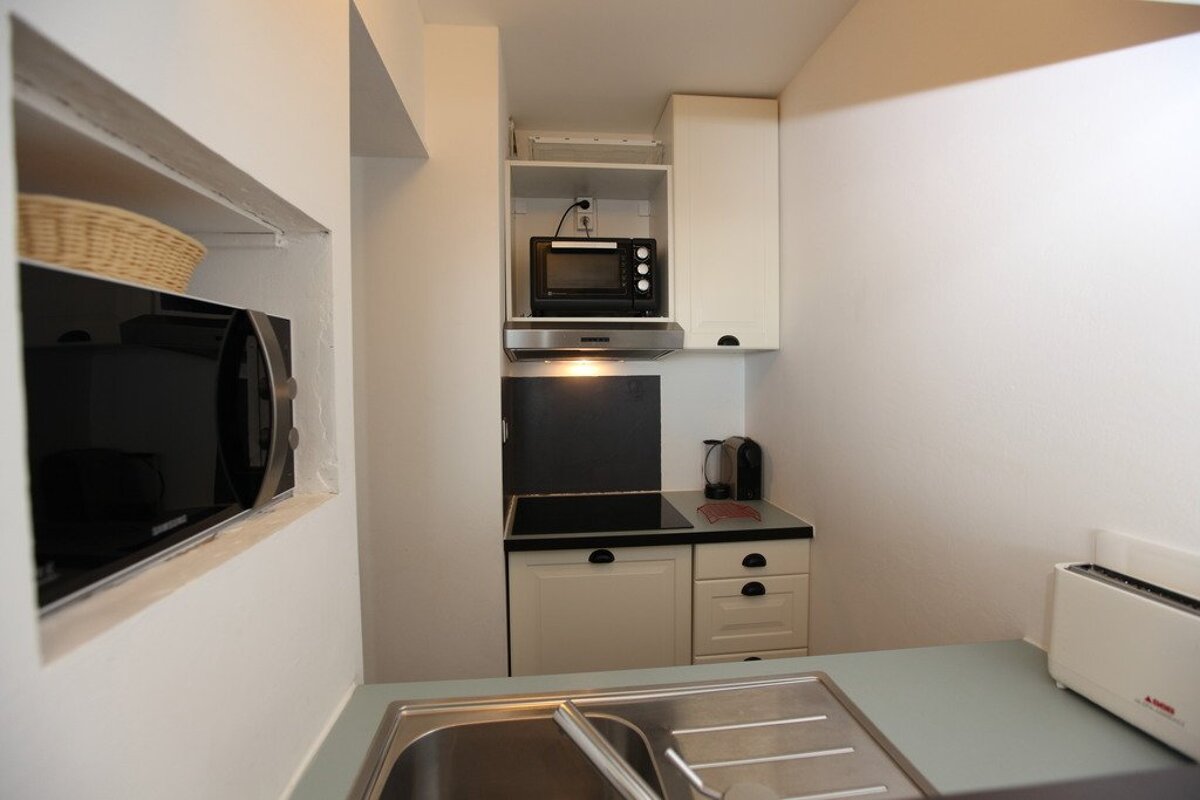 A compact, L-shaped kitchen with a sink, built-in microwave, a small oven over a hob, and a toaster. It has white walls and cream cabinets.