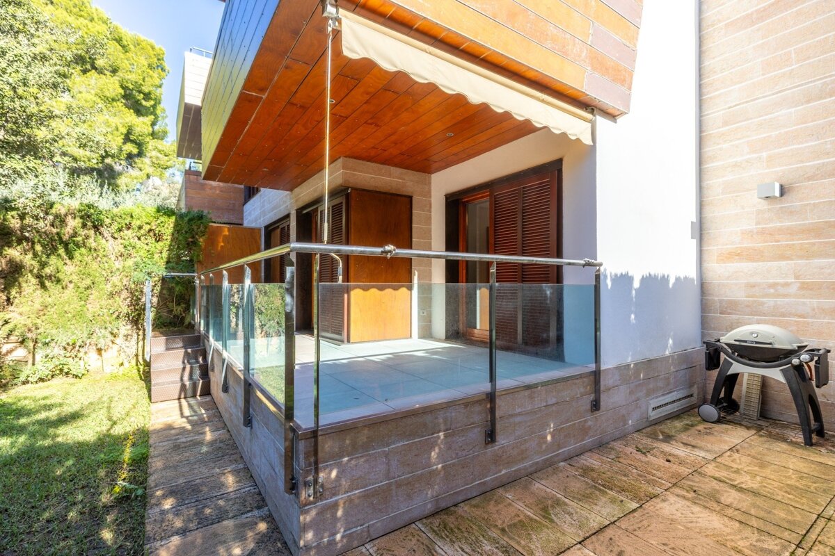 A modern outdoor patio with a glass railing, wooden ceiling, and a BBQ grill, next to a lush green garden and house facade.