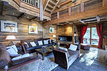 A spacious, rustic log cabin living room featuring warm wood, plush sofas, a grand stone fireplace, and a picturesque snowy landscape seen through red curtains.