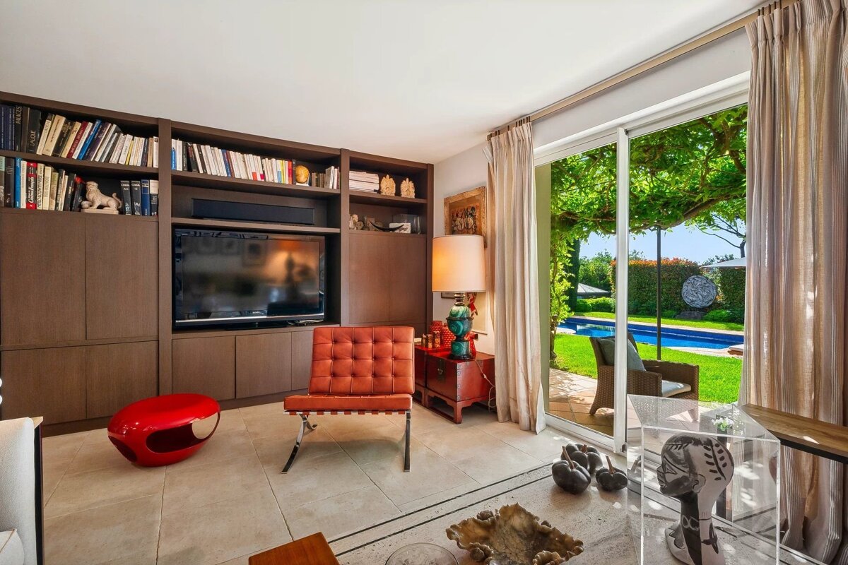Cozy living room with a large built-in bookshelf, orange leather chair, and glass doors opening to a scenic backyard pool.