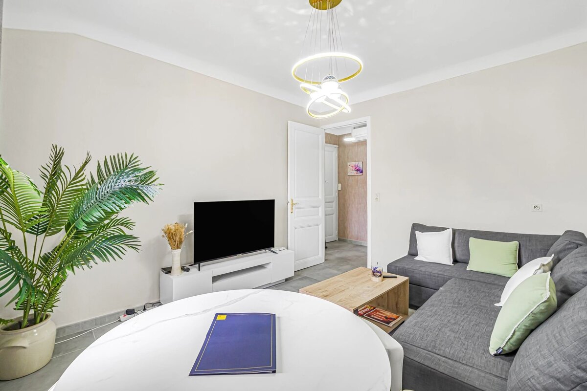 Bright modern living room with a grey sectional sofa, white round dining table, TV, wooden coffee table, and a large potted plant.