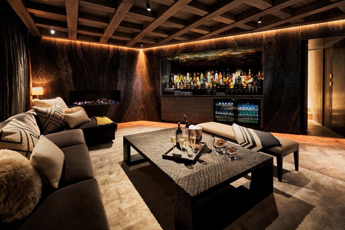 A warm, upscale lounge features dark wood walls, a glowing bar, fireplace, plush seating, and drinks on a textured coffee table.