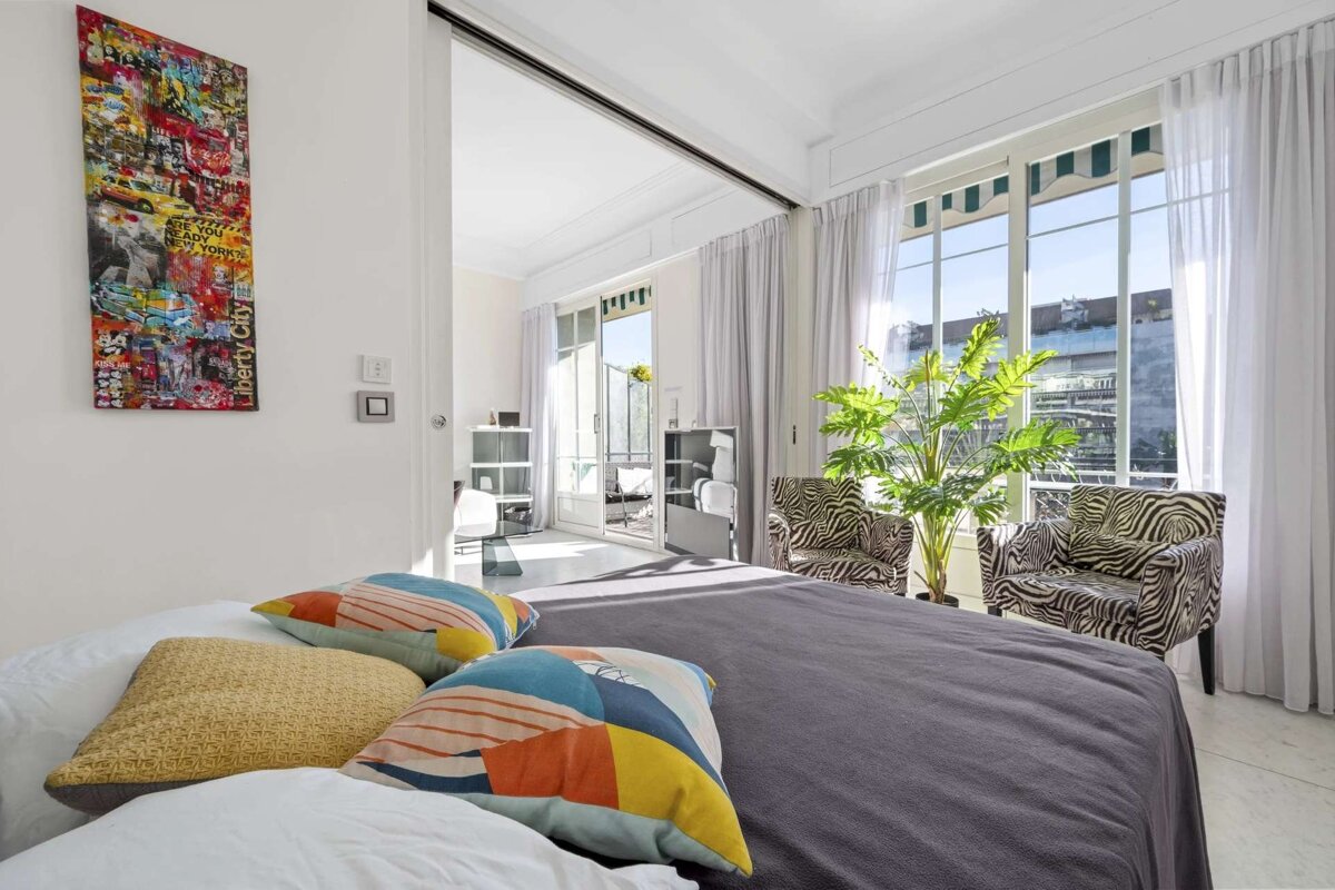 Bright bedroom with a bed, colorful pillows, zebra chairs, a large plant, and artwork. Large windows open to a balcony/terrace.