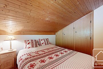 Cozy alpine-style bedroom with a sloped wooden ceiling, a bed adorned with festive red and white reindeer/snowflake bedding, and built-in wood wardrobes.