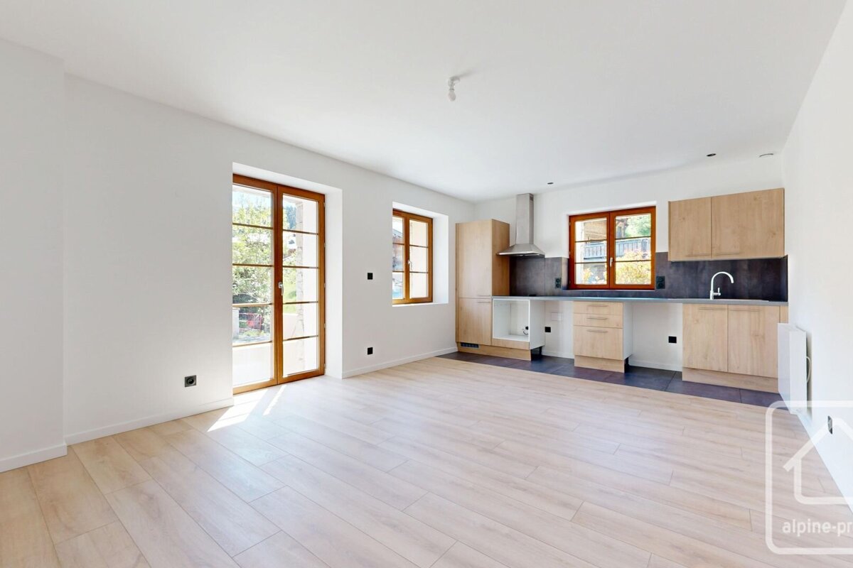Bright, modern open-plan room with a light wood kitchen area, light wooden floors, white walls, and large glass doors/windows providing ample light.