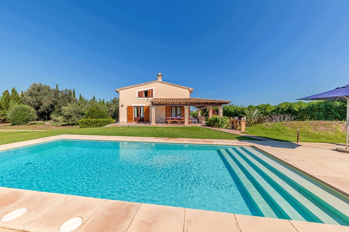 A light brown house with a covered patio, a sparkling blue swimming pool, and a lush green garden under a clear blue sky.