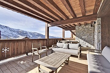 A wooden balcony with a couch and chairs and mountains in the background