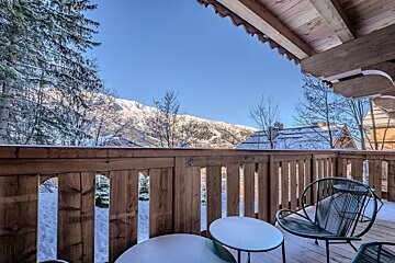 A wooden chalet balcony with modern outdoor chairs overlooks a stunning, snow-covered mountain landscape under a clear blue sky.