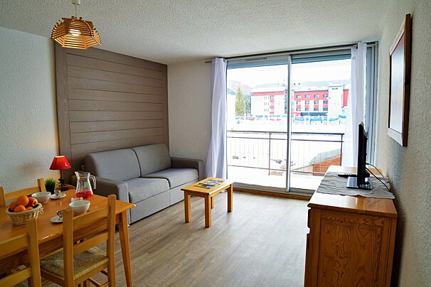 A bright living/dining room with a grey sofa, wooden dining set, TV, and large sliding doors opening to a balcony with buildings outside.