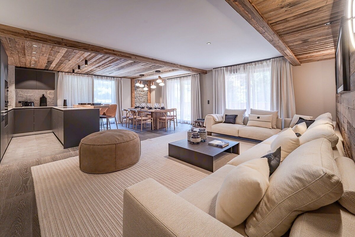 A modern open-plan living space with a large sofa, kitchen island, dining table, and rustic wooden ceilings.