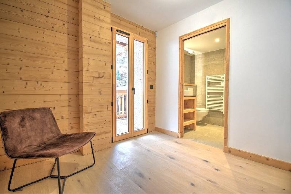 A room with wood-paneled walls, light wood floor, brown chair, glass doors to a balcony, and an open doorway to a bathroom.