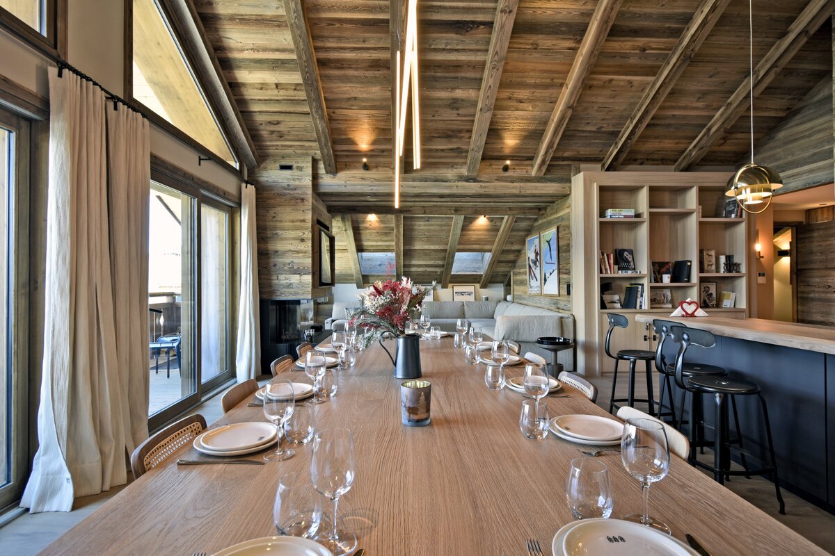 An elegant ski chalet's open-plan dining and living space, featuring a long wooden table, exposed beams, large windows, and a sophisticated bar area.