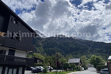 A dark wooden chalet in a mountain village, backed by snow-capped peaks, green forests, under a partially cloudy sky.