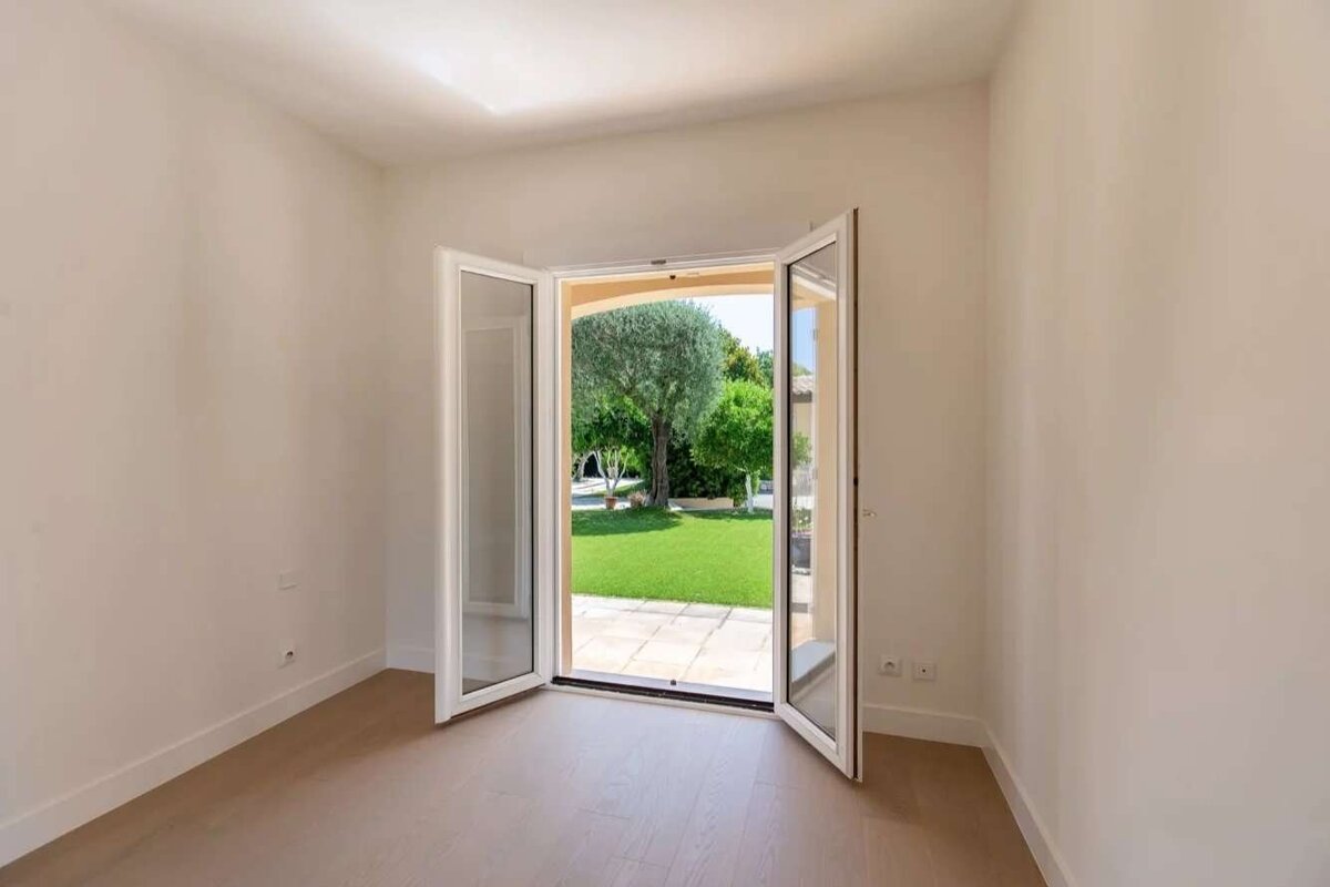 An empty room with light walls and wood floor, featuring open double glass doors leading to a lush green garden with a large tree and stone patio.