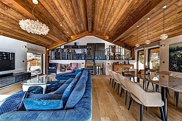 Spacious, modern open-concept chalet interior with vaulted wood ceiling, blue sofa, dining table, and a contemporary kitchen on a mezzanine.