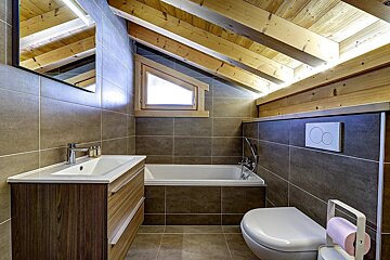 A modern attic bathroom featuring a slanted wooden beamed ceiling, brown tiled walls, a bathtub, toilet, and vanity with sink, plus a triangular window.