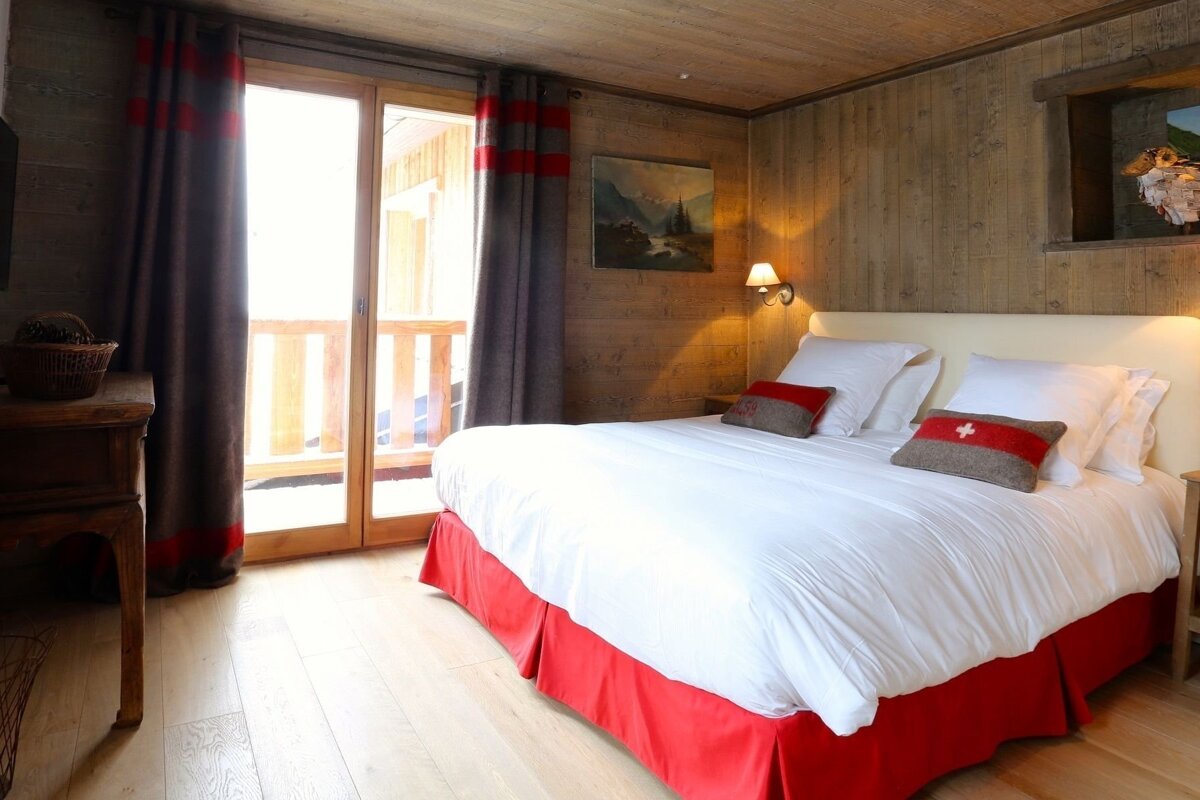 A cozy chalet-style bedroom with wood walls, a white bed with red skirt & Swiss flag pillows, and a bright balcony door.