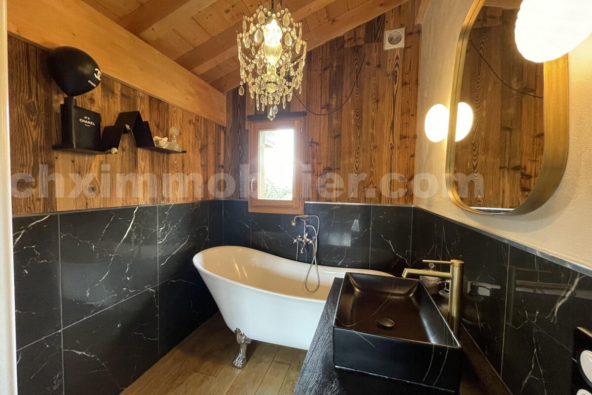 A luxurious rustic bathroom features wood paneling, black marble walls, a white clawfoot tub, and a black vessel sink, illuminated by a chandelier.