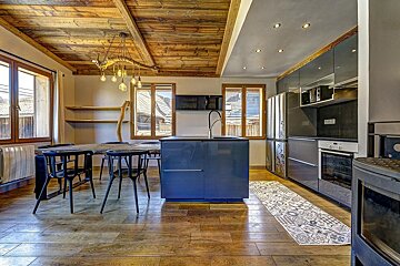 A modern kitchen and dining area with a rustic wooden ceiling and floor, featuring a blue island, sleek grey cabinets, and a large dining table.