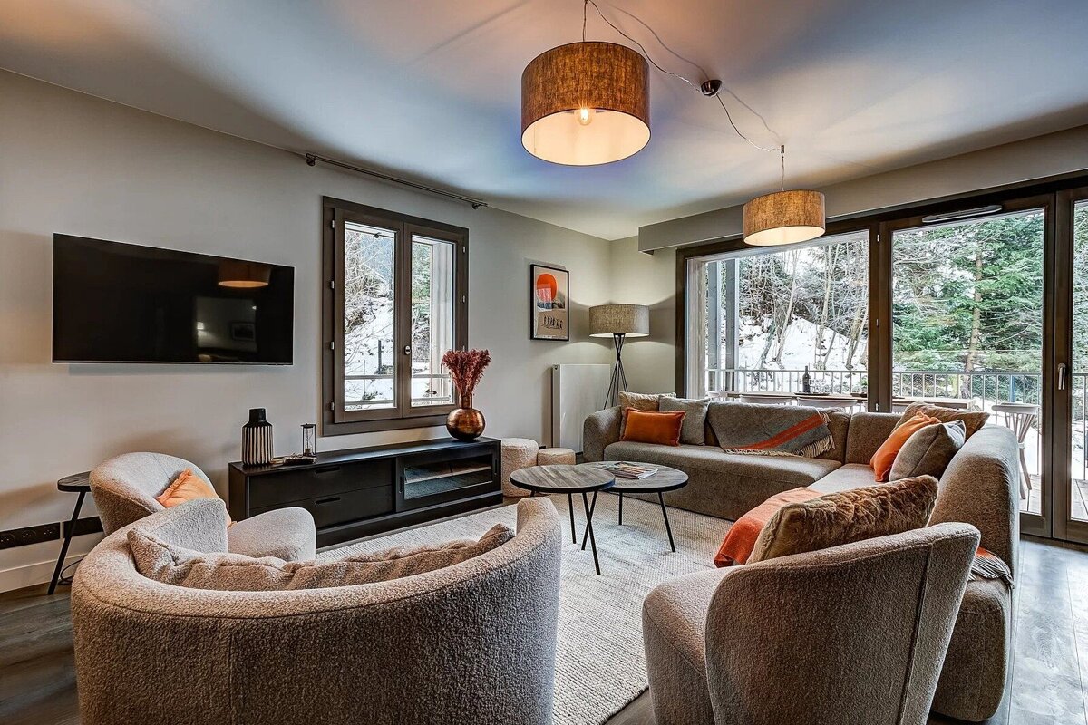 Cozy, modern living room featuring plush curved armchairs, a sectional sofa, and large windows looking out to a snowy forest.
