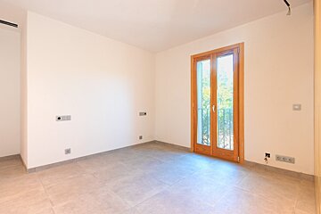 An empty, clean room with light walls and a tiled floor. A large double wooden door with glass panels leads to a balcony with railings.