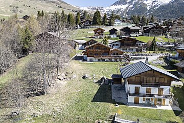 An alpine village with wooden chalets on a grassy hillside, backdropped by snow-capped mountains.