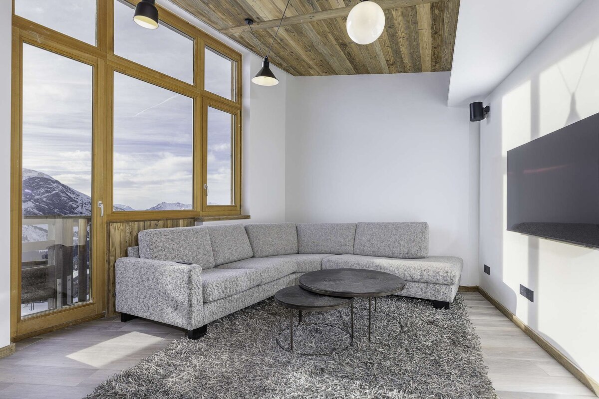 Modern living room with a grey sectional sofa, nested coffee tables, wooden ceiling, and large window overlooking snowy mountains.