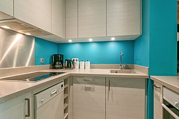 A modern kitchen with bright turquoise walls, light wood cabinets, and light gray counters. It includes a stovetop, sink, dishwasher, coffee maker, and kettle.