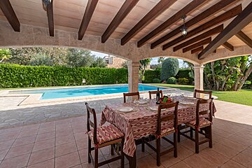 A table and chairs under a covered patio with a pool in the background