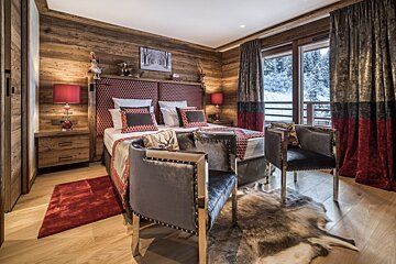 A luxurious chalet bedroom with wood paneling, a large bed with red accents, two velvet chairs, and a picturesque view of a snowy forest.