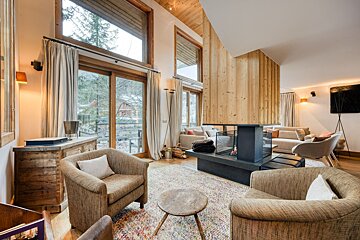 A cozy modern chalet living room featuring a central fireplace, large windows with a view, and comfortable seating amidst warm wood accents.