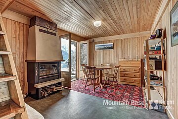 Rustic, wood-clad chalet interior with a cozy fireplace, small dining set on a red rug, and a doorway revealing a snowy mountain view.