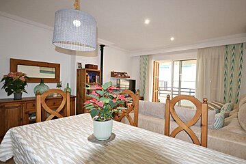 A bright, open-plan dining and living room featuring wooden furniture, plants, a patterned tablecloth, and a balcony view.