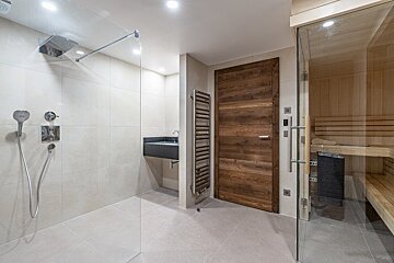 Modern spa bathroom with walk-in shower, dark wood door, and glass-front sauna. Features a sink and towel warmer.