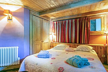 Cozy rustic bedroom featuring a wooden ceiling, a double bed with folded bathrobes, striped curtains, and warm lighting.