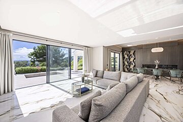 Spacious, modern living room with a gray sectional, marble floors, and large glass doors opening to a lush landscape. A stylish dining area is visible.