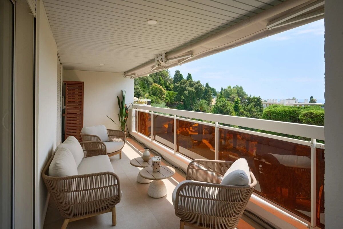 Modern apartment balcony with stylish wicker seating, an amber glass railing, and a beautiful view of lush green trees.