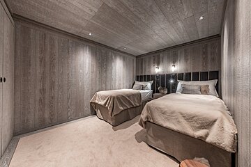 A cozy twin bedroom featuring wood-paneled walls and ceiling, soft carpet, plush beds with dark headboards, and warm accent lighting.