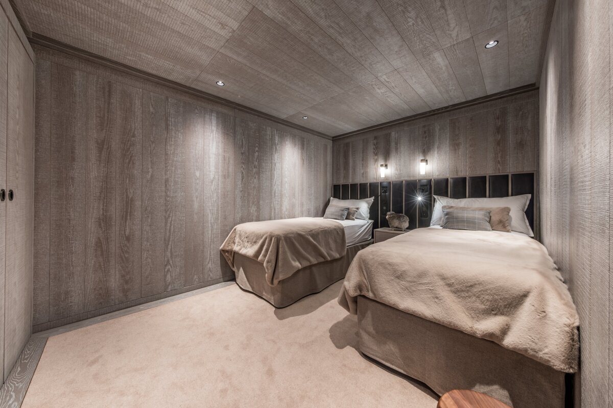 A cozy twin bedroom with wood-paneled walls and ceiling, neutral bedding, and a soft carpet. Warm lighting enhances the inviting atmosphere.