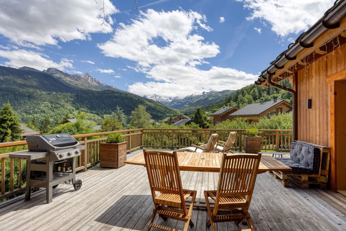 Rustic wooden deck with a grill and dining set, offering breathtaking views of lush green mountains, some snow-capped, under a vibrant blue sky.