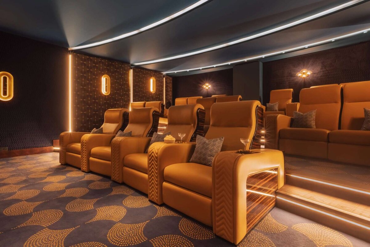 A high-end home cinema with mustard yellow recliners, a dark patterned carpet, glowing accent lights on walls and steps, and a curved ceiling.