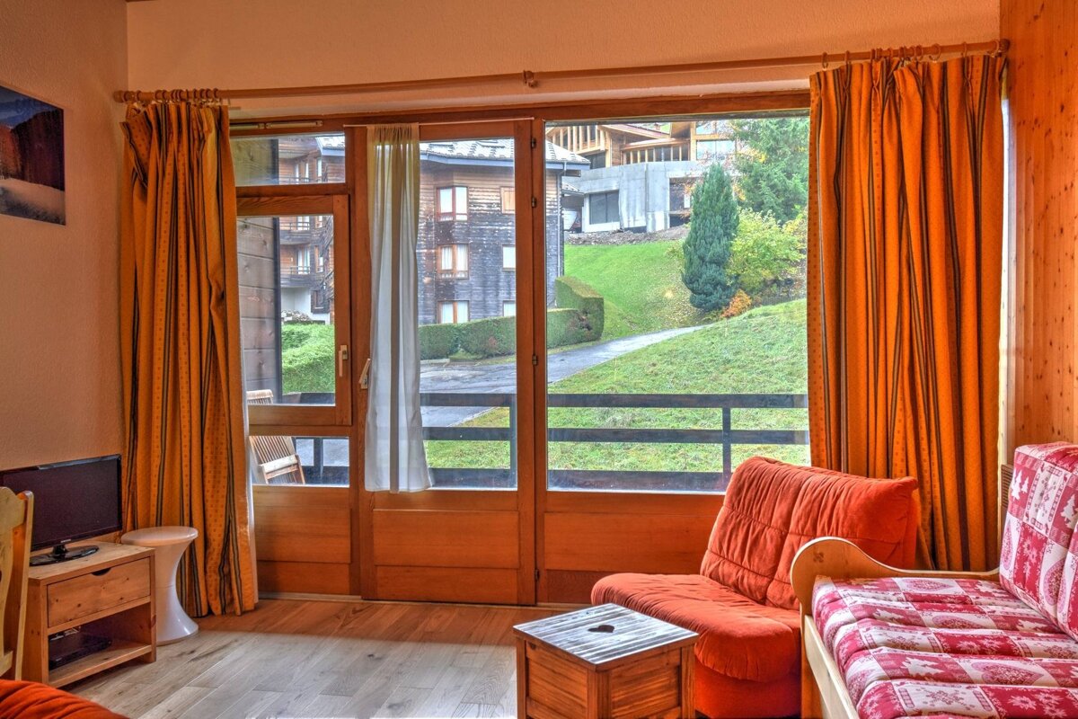 A cozy room with orange curtains and furniture, featuring a large window overlooking a green, hilly landscape with distant buildings.
